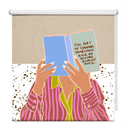 A decorative window roller shade featuring a colorful graphic illustration of a person in pink striped pajamas holding a blue book with a quote on the cover, set against a beige and white background with brown speckles.