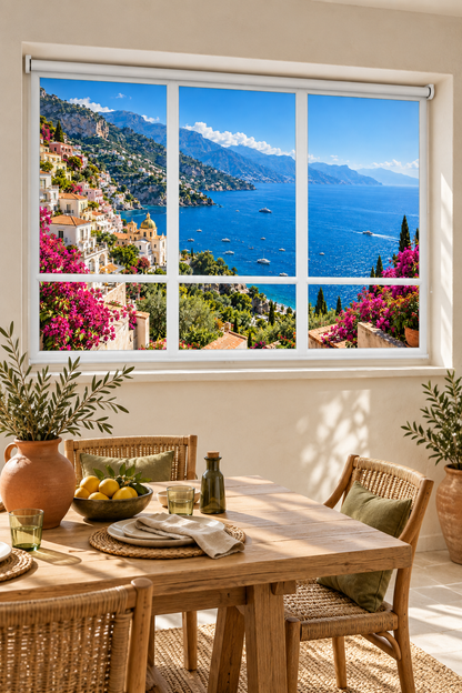 A large wall window or mural showing a Mediterranean coastal town with blue sea, mountains, and pink flowers, installed above a wooden dining table with chairs and plants in a bright room.