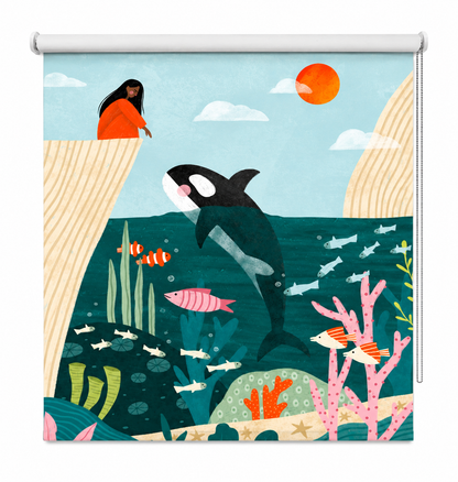 A stylized illustrated ocean scene printed on a window roller shade, showing an orca, fish, coral, a person in an orange dress on a cliff, clouds, and a sun in a flat, colorful art style.