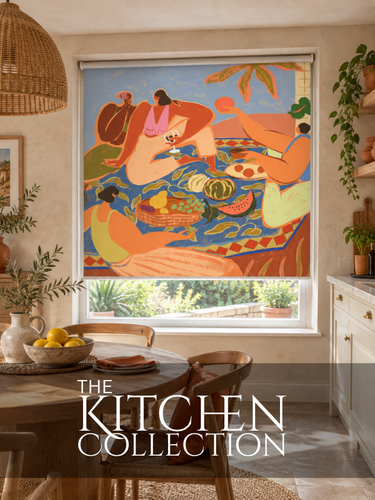A colorful roller blind featuring a stylized painting of women at a picnic, installed on a window in a warm, modern kitchen setting. A dark banner at the bottom reads 'THE KITCHEN COLLECTION'.