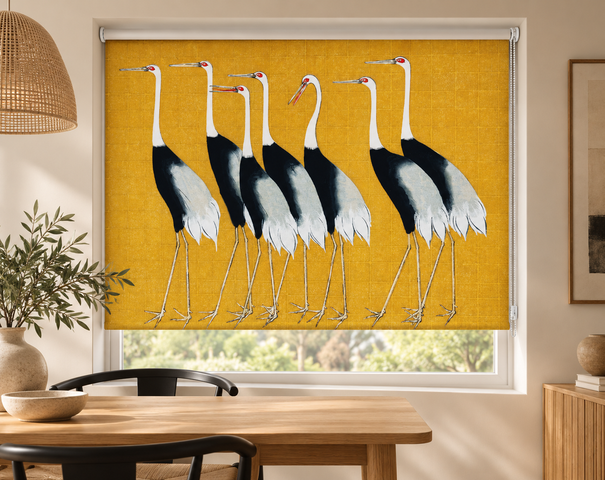 A window roller shade featuring a printed design of seven storks on a mustard yellow background, shown installed in a modern dining room setting.