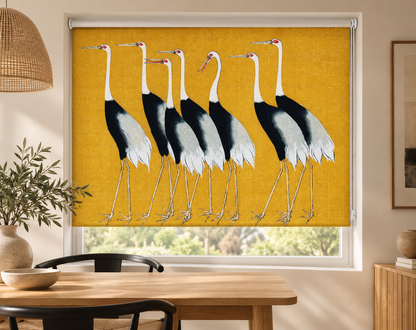 A window roller shade featuring a printed design of seven storks on a mustard yellow background, shown installed in a modern dining room setting.